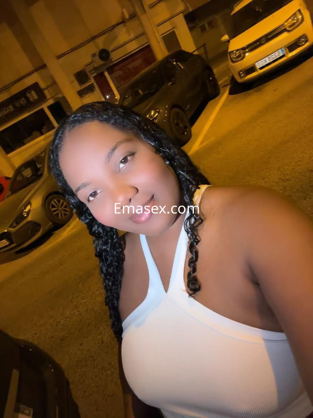 Escort en Tarragona posando con un top blanco, cabello trenzado, en un entorno urbano iluminado por la noche.