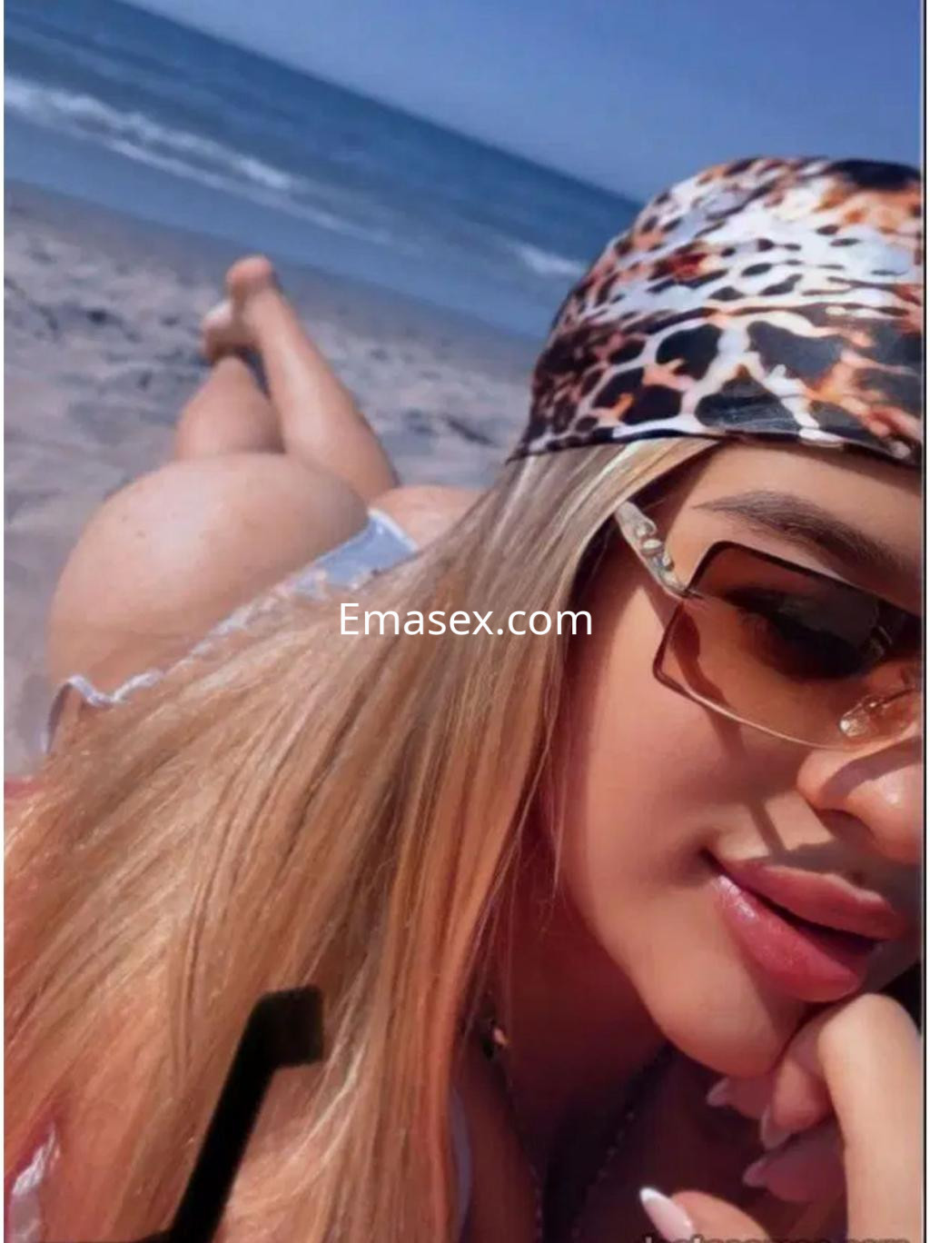 Puta en Irún con cabello largo y rubio, usando gafas de sol y un pañuelo de animal print, relajándose en la playa.
