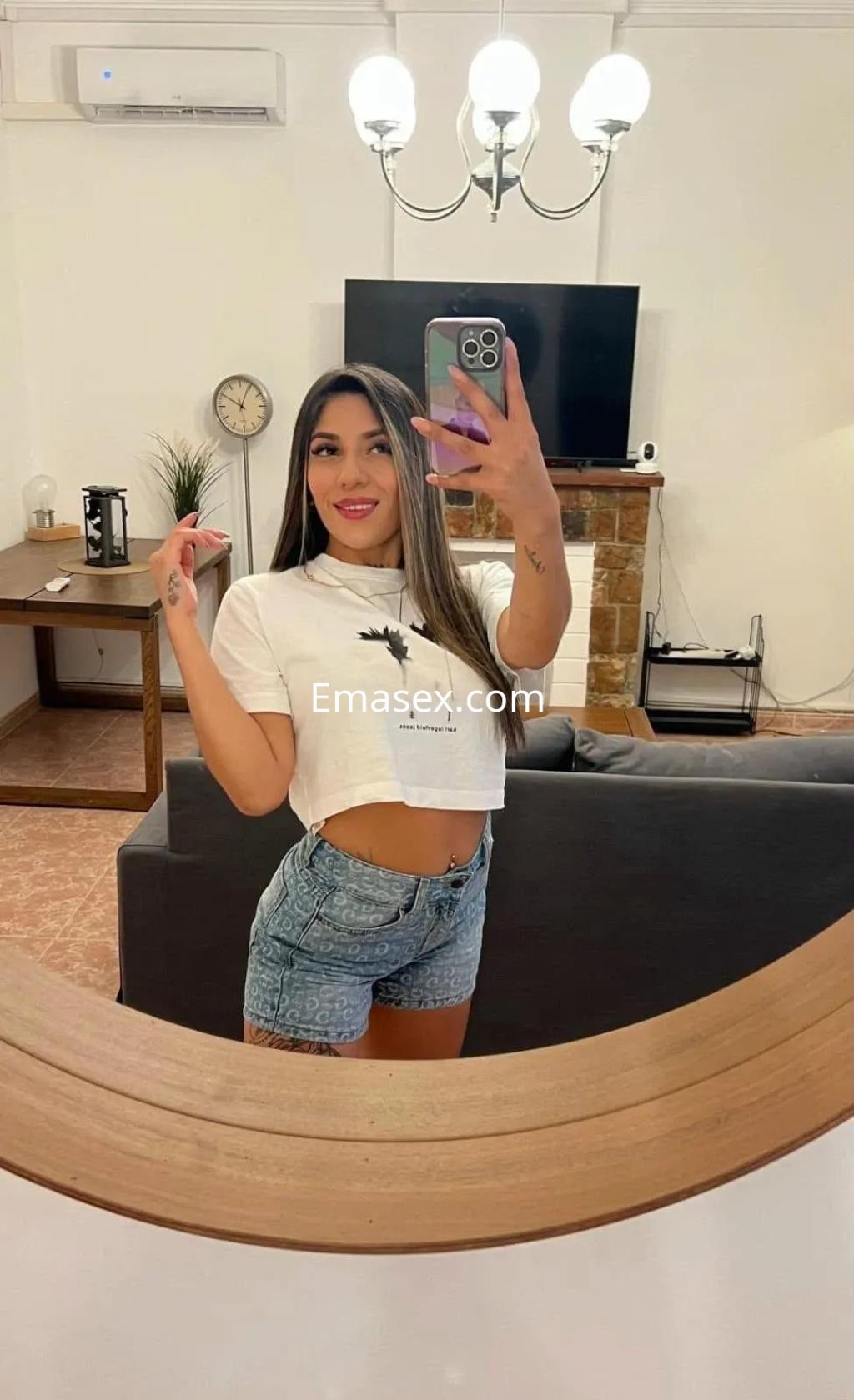 Puta en Palma de Mallorca, con camiseta corta y shorts de mezclilla, posando frente a un espejo en un salón.