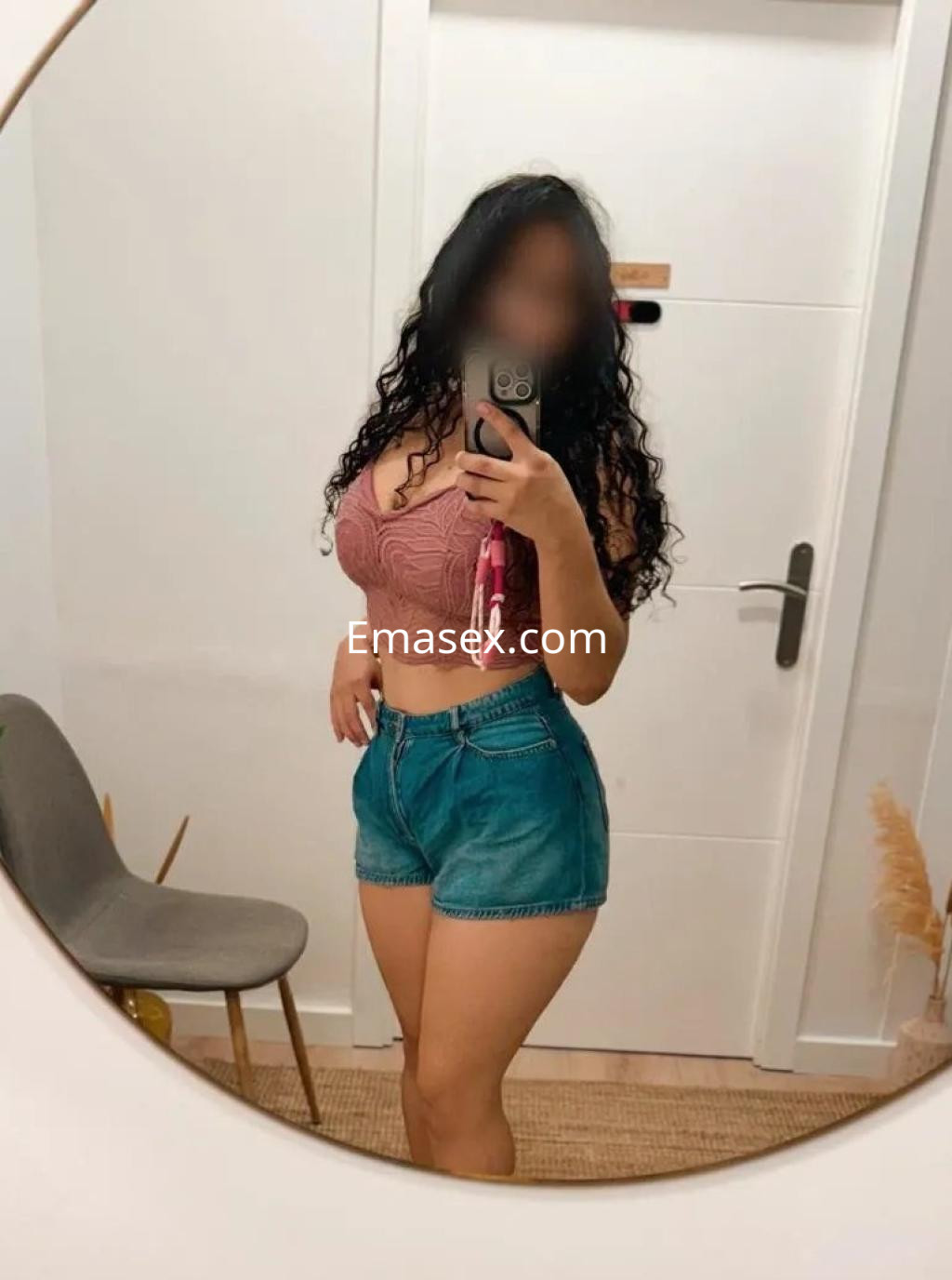 Escort con cabello rizado y oscuro, viste un top rosa y shorts de mezclilla, posando frente a un espejo en Palma de Mallorca.