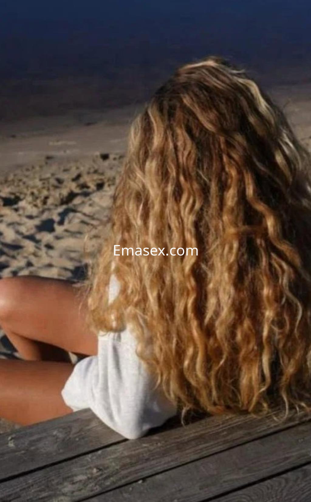 Puta en Las Palmas sentada en la playa, con cabello rizado y largo, vistiendo una camiseta clara.