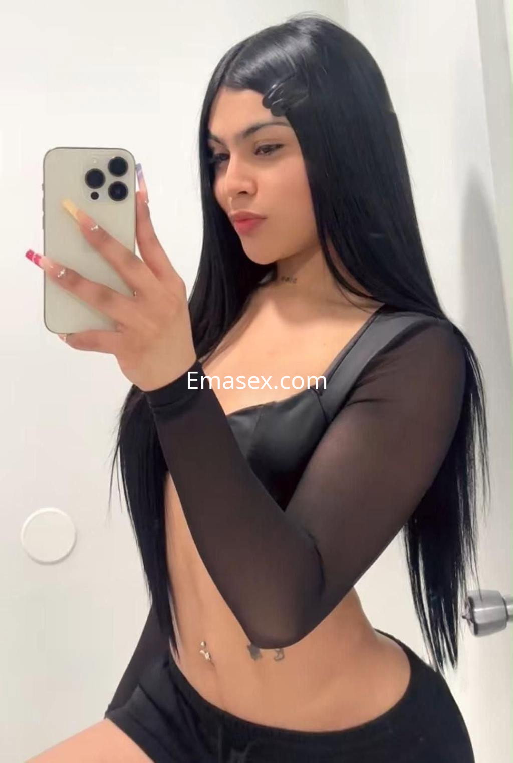 Escort en Granada con cabello largo y lacio, vistiendo una blusa negra transparente y shorts oscuros, posando con un espejo.