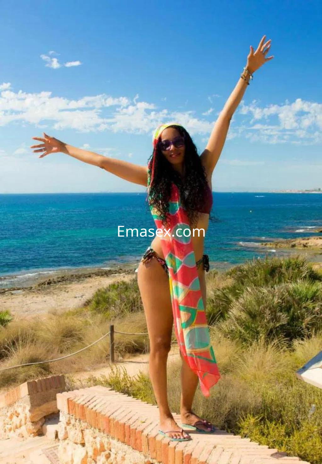 Puta con cabello rizado y colorido, luciendo un bikini y pareo en la playa de Alicante, con brazos en alto.