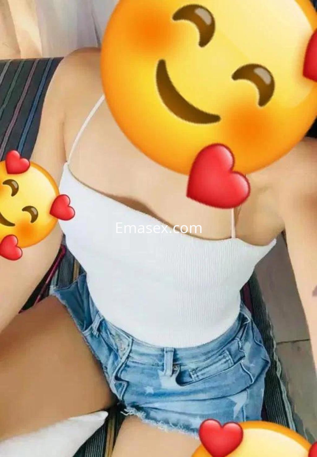 Escort en Alicante sentada, vistiendo un top blanco ajustado y shorts de mezclilla, con fondo de rayas.