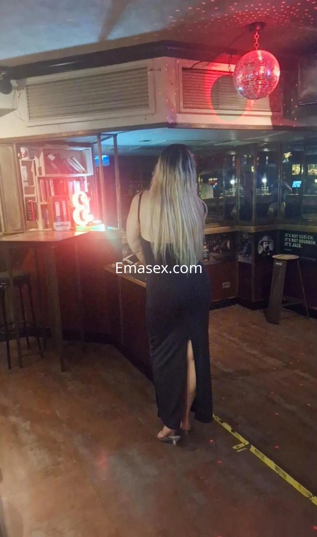 Escort de espaldas en un bar de Santander, luciendo un vestido largo negro con una abertura en la pierna.
