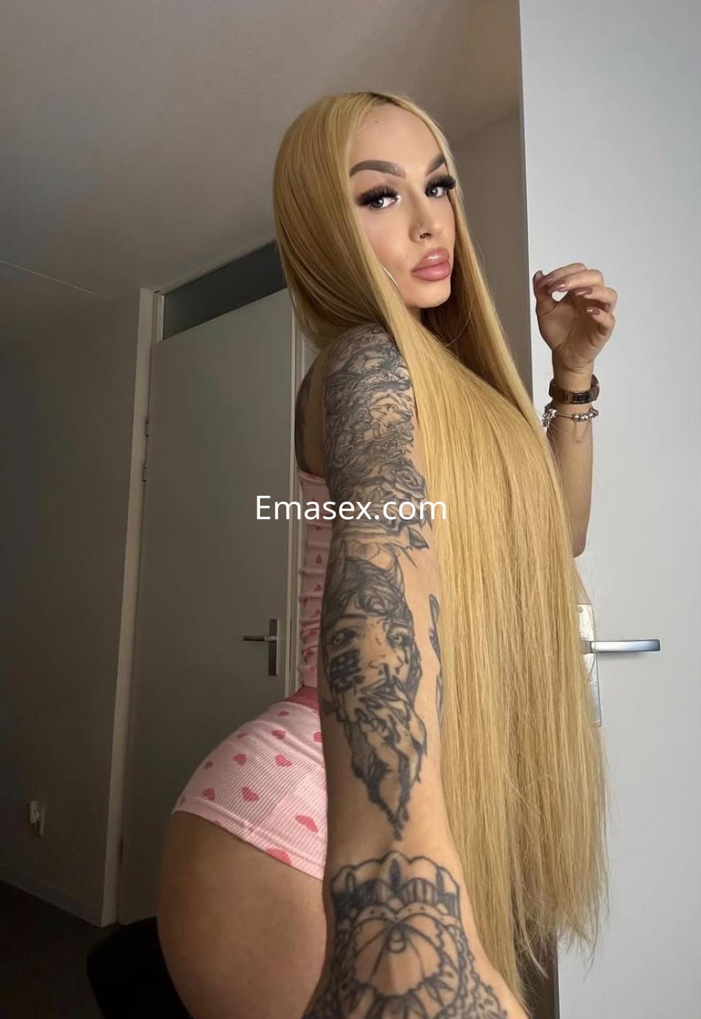 Escort trans en Pamplona, con cabello largo y rubio, luciendo un conjunto de lencería rosa con corazones y tatuajes visibles.