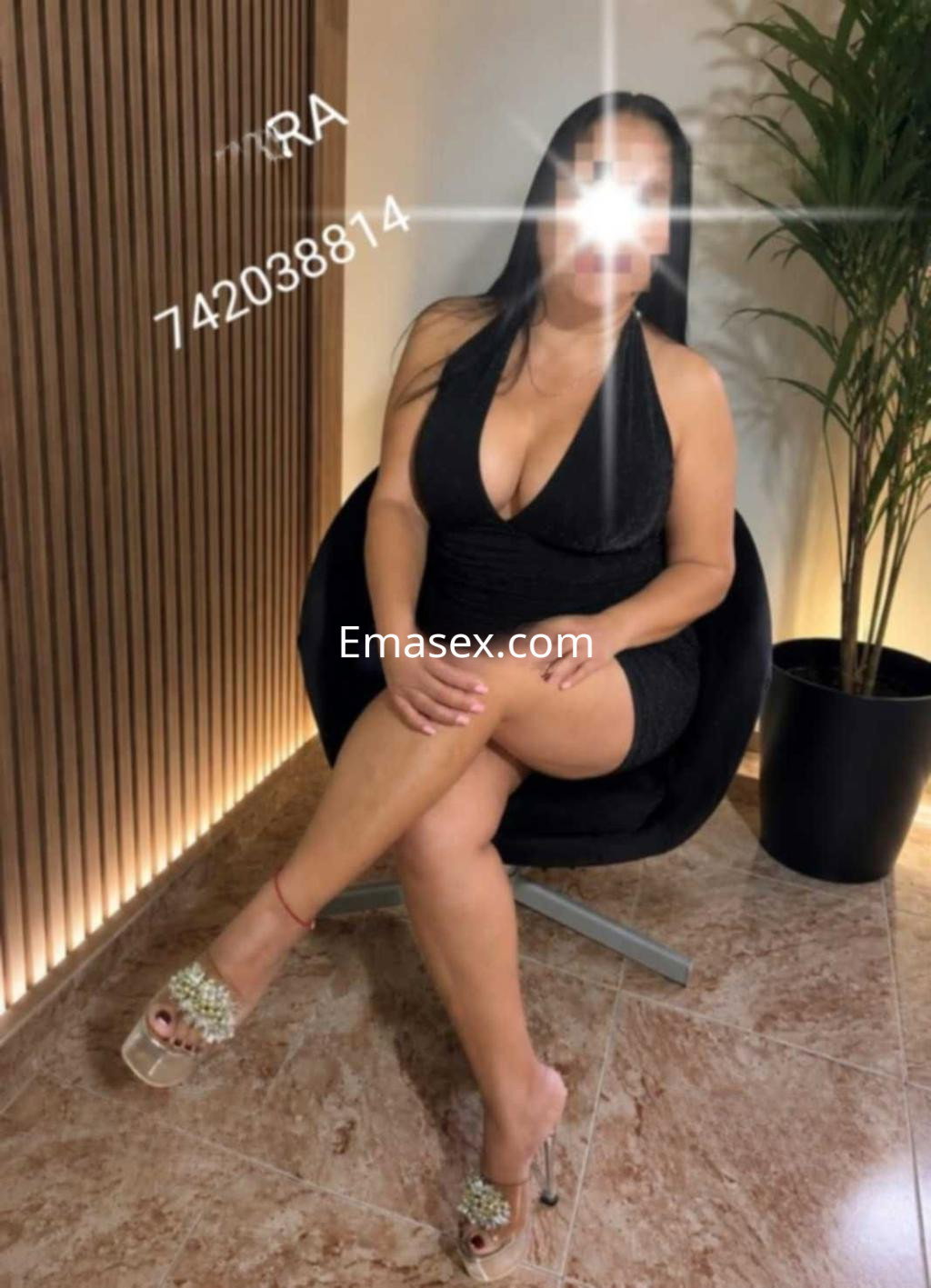 Escort en Zamora, sentada en una silla negra, luciendo un vestido ajustado y sandalias brillantes.