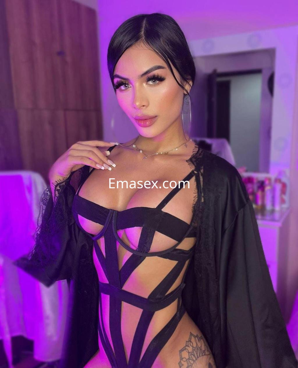 Puta en Pamplona lleva un body negro con tiras y una bata, posando con cabello lacio y un fondo iluminado.