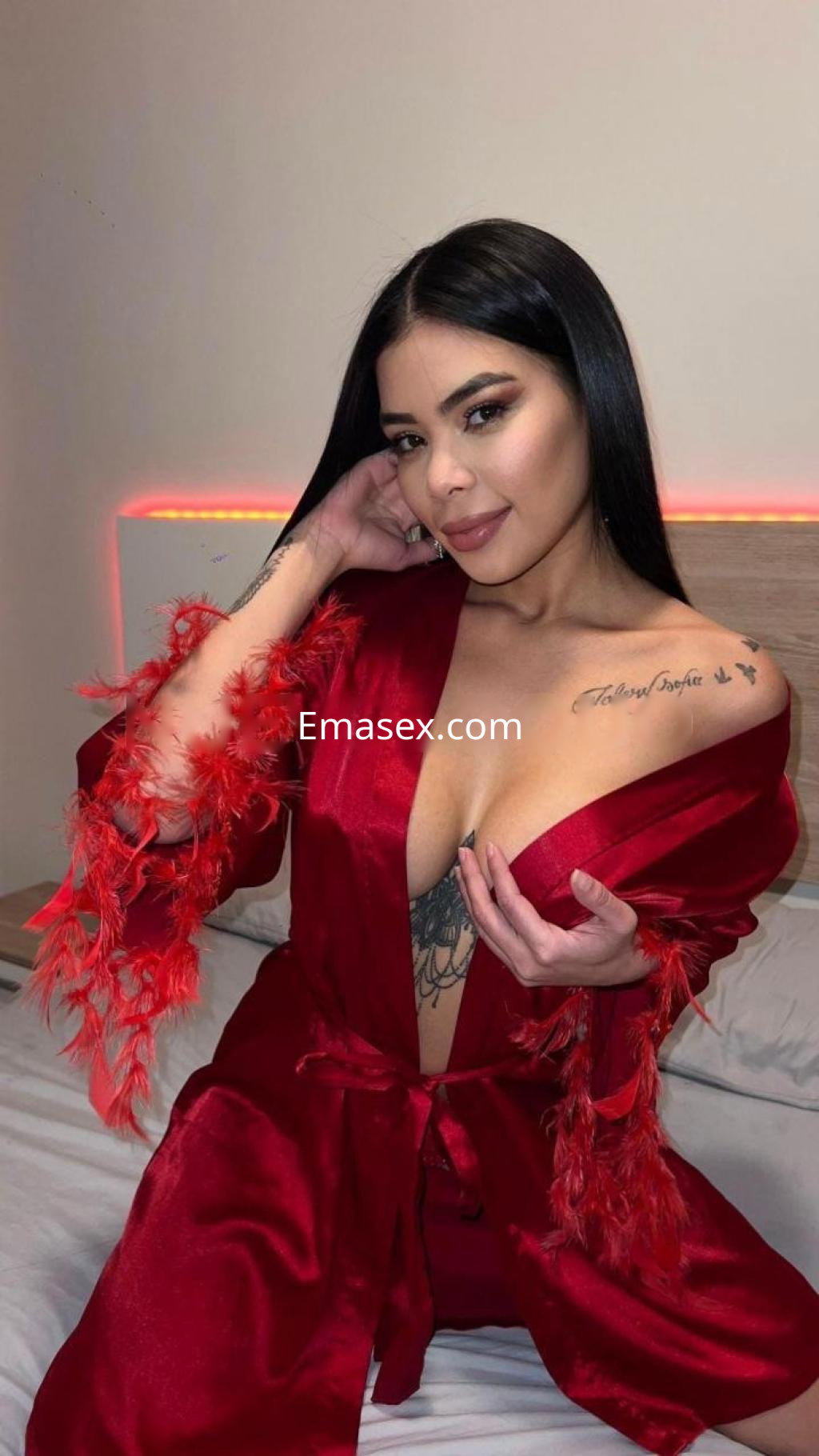 Puta en Barcelona viste un robe rojo con detalles de plumas, mientras posa en una cama con un fondo iluminado.