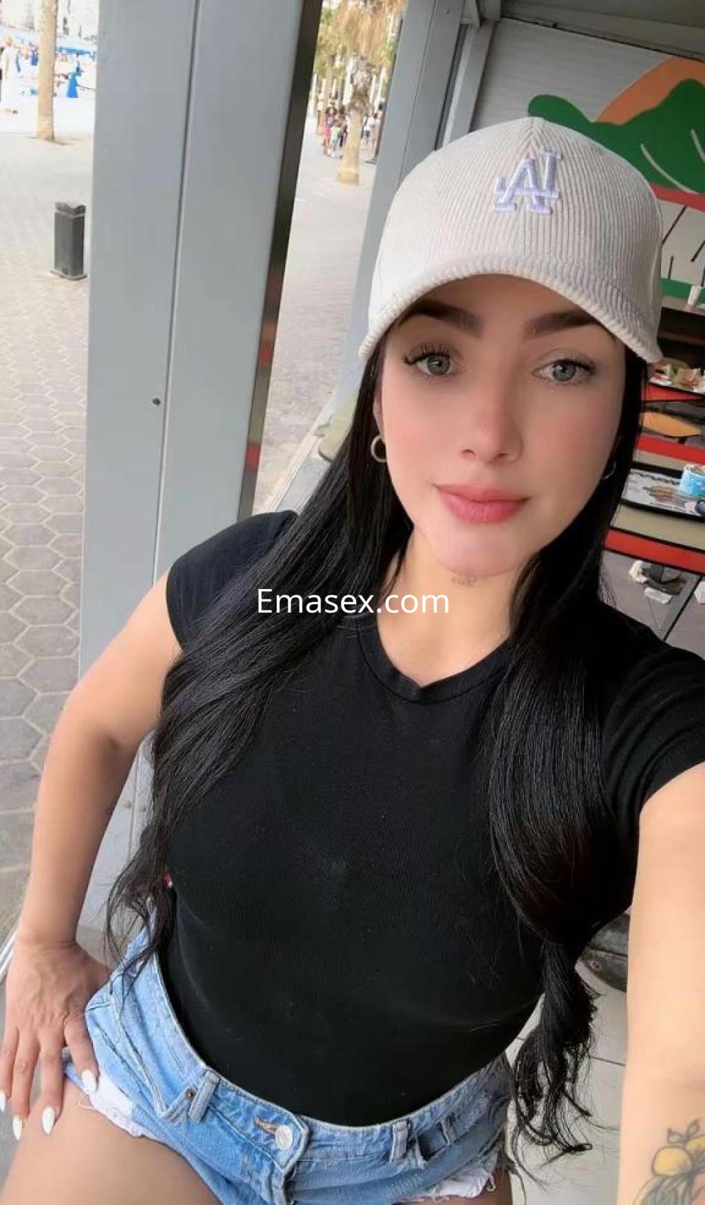 Puta en Alicante con cabello largo y lacio, usando una gorra y una camiseta negra, sentada en un entorno al aire libre.