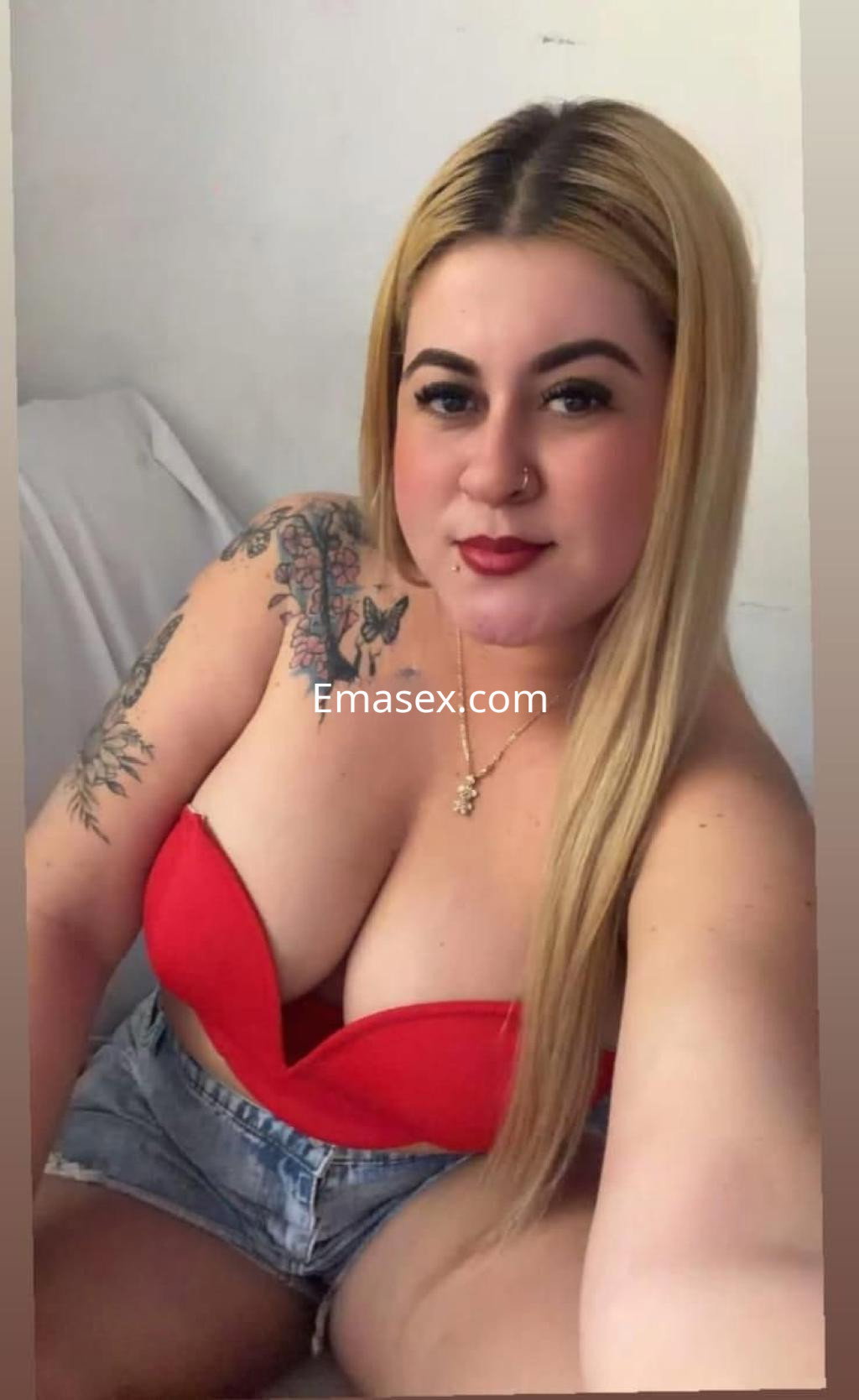 Escort en León posando con blusa roja y shorts de mezclilla, mostrando tatuajes y cabello largo y rubio.