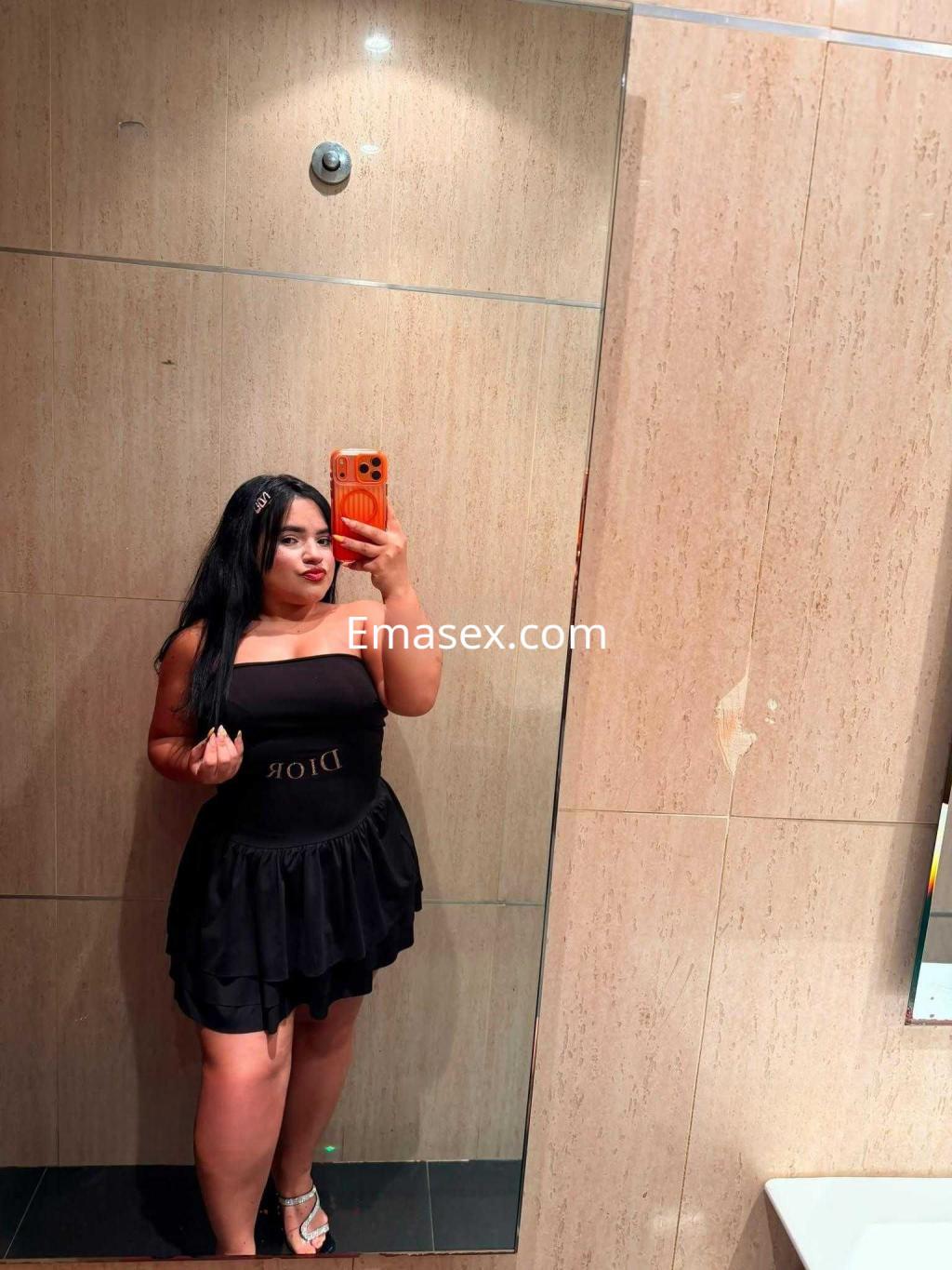 Puta posando frente a un espejo en un vestido negro sin mangas con logo de Dior, en un entorno moderno en Murcia.