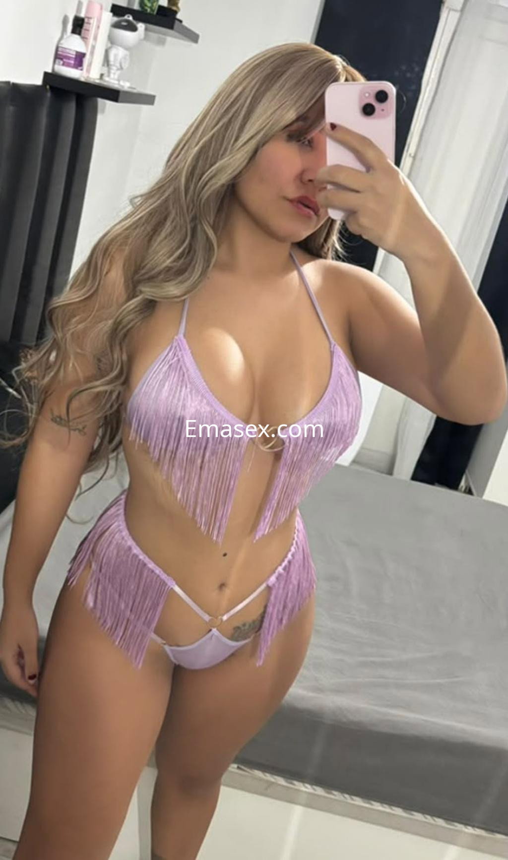 Escort en Alicante con cabello largo y rubio, vistiendo un conjunto de lencería rosa con flecos, posando frente al espejo.