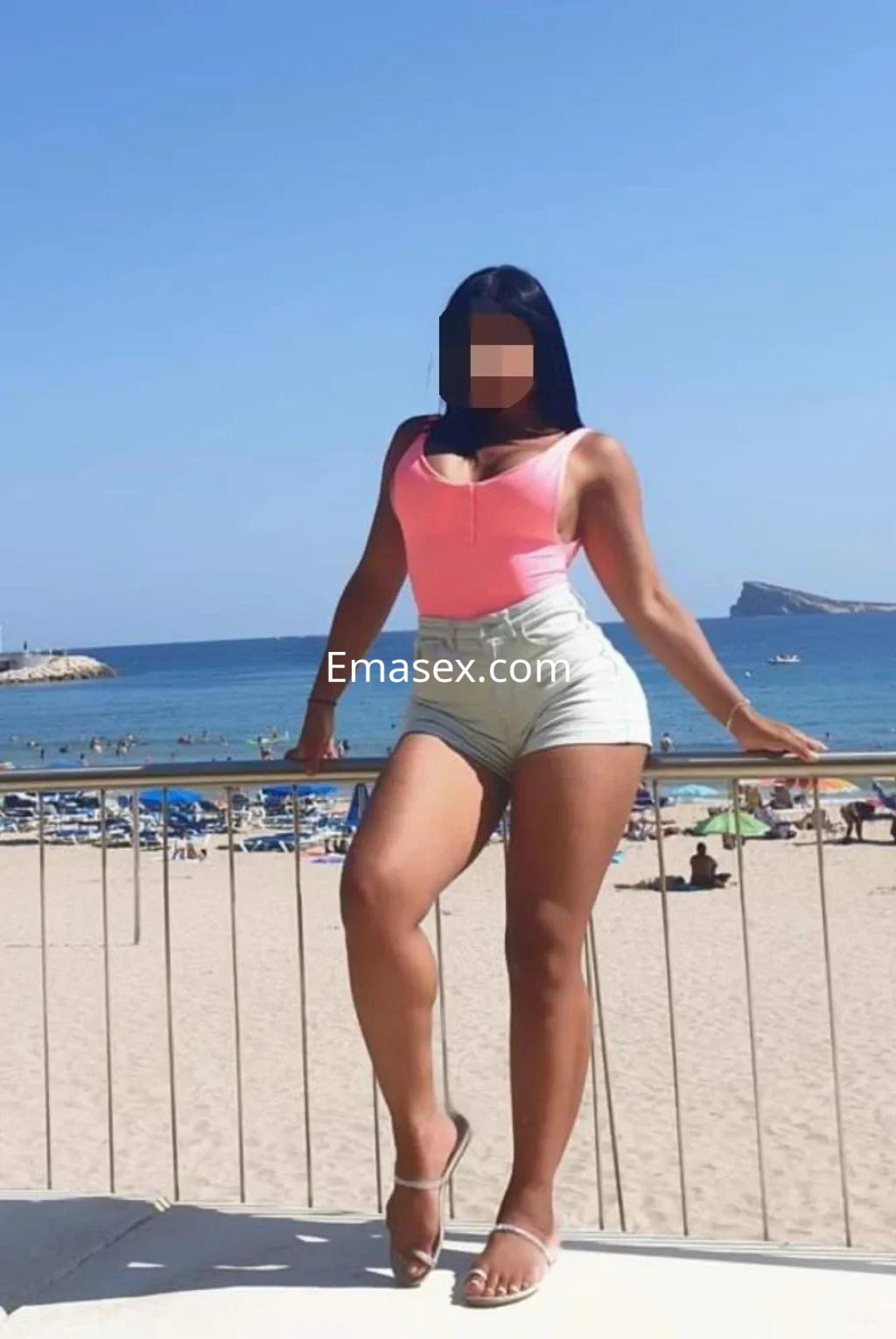 Escort en Vitoria, posando al aire libre junto a la playa, con blusa rosa y shorts claros.