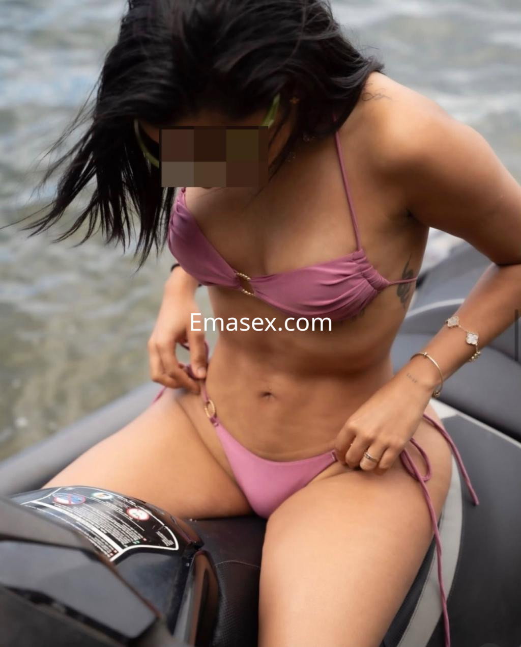 Escort en Huelva luciendo un bikini rosa mientras ajusta su ropa en una moto de agua cerca del mar.