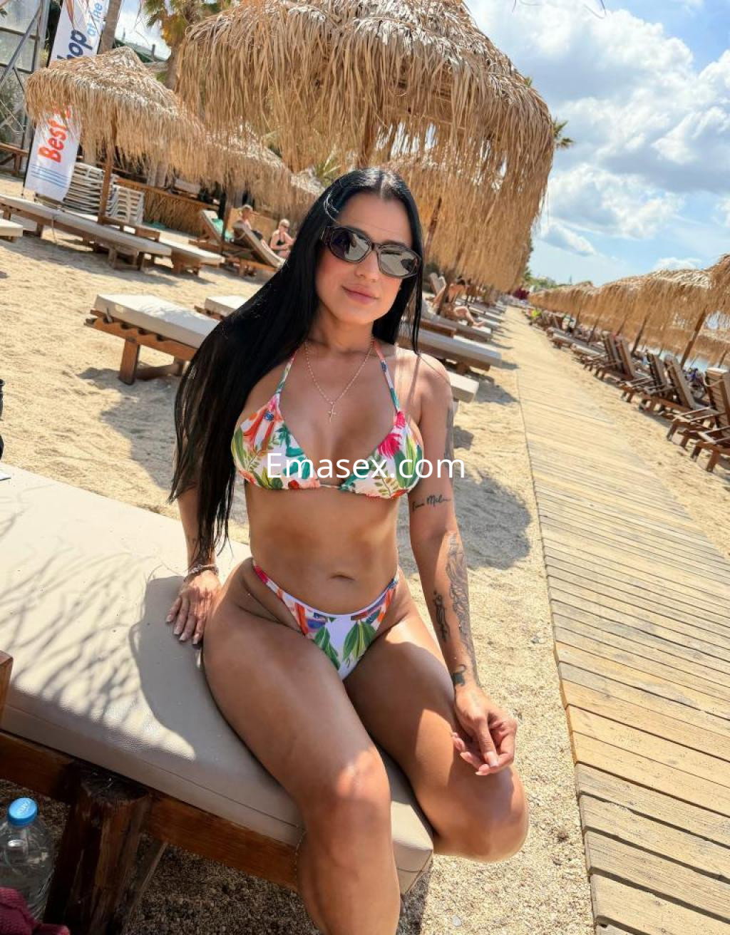 Escort en Murcia, luciendo un bikini colorido mientras se relaja en una playa con tumbonas y sombrillas de paja.
