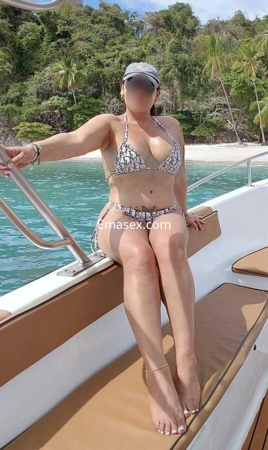Escort en Murcia, posando en un yate con un bikini de estampado de serpiente y una gorra, junto al mar.