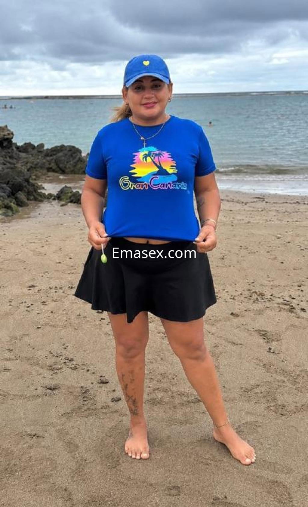 Escort en Las Palmas vistiendo una camiseta azul con el logo de Gran Canaria y una falda negra en la playa.