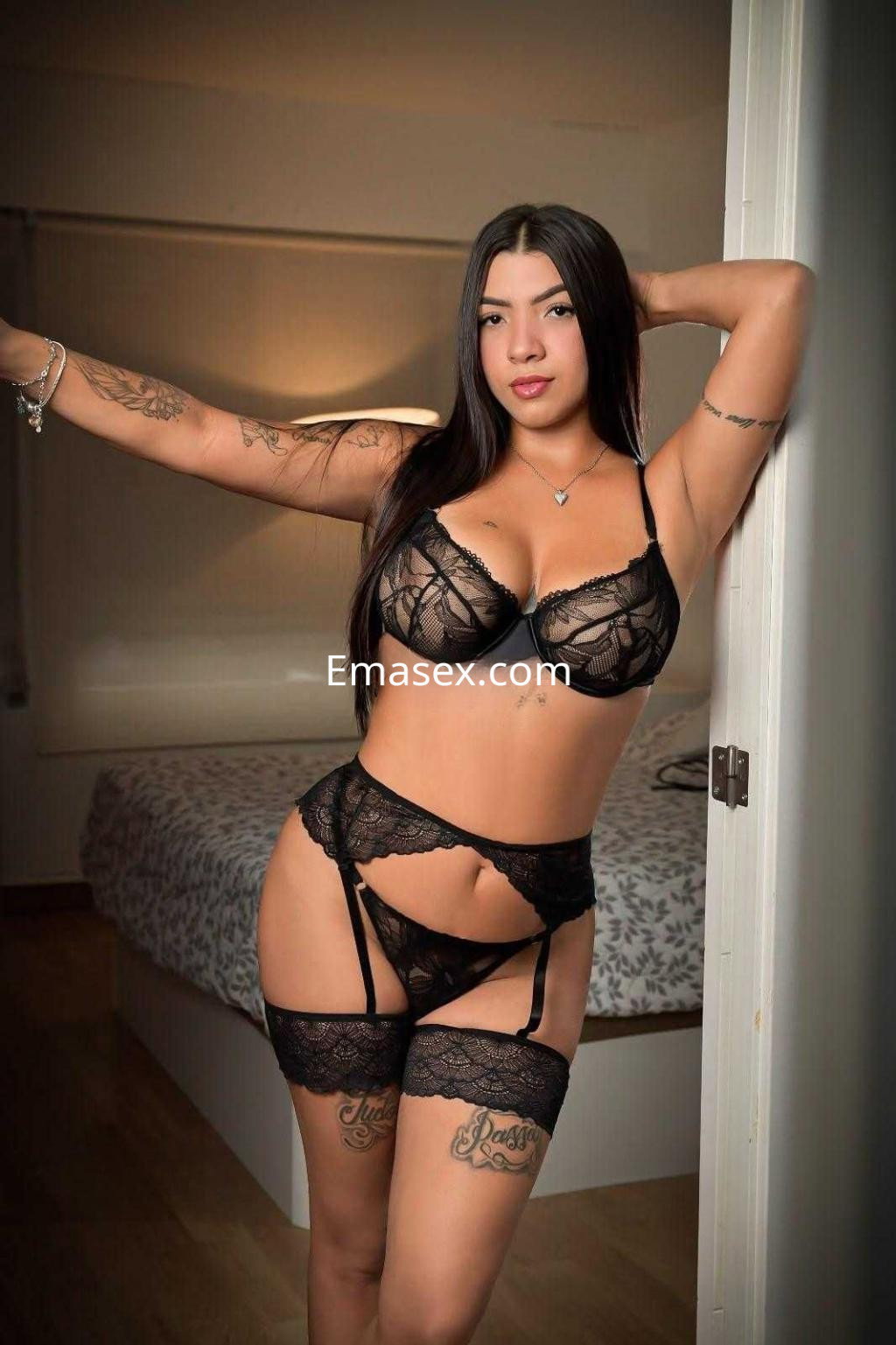 Puta en Alicante luce lencería negra con encaje, posando de pie en un ambiente interior con cama al fondo.