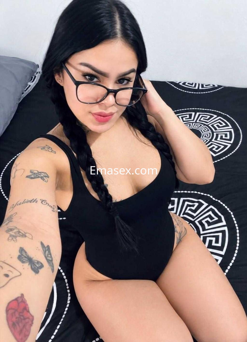 Escort en Zaragoza con cabello negro trenzado, vistiendo un body negro y gafas, tomando una selfie en la cama.