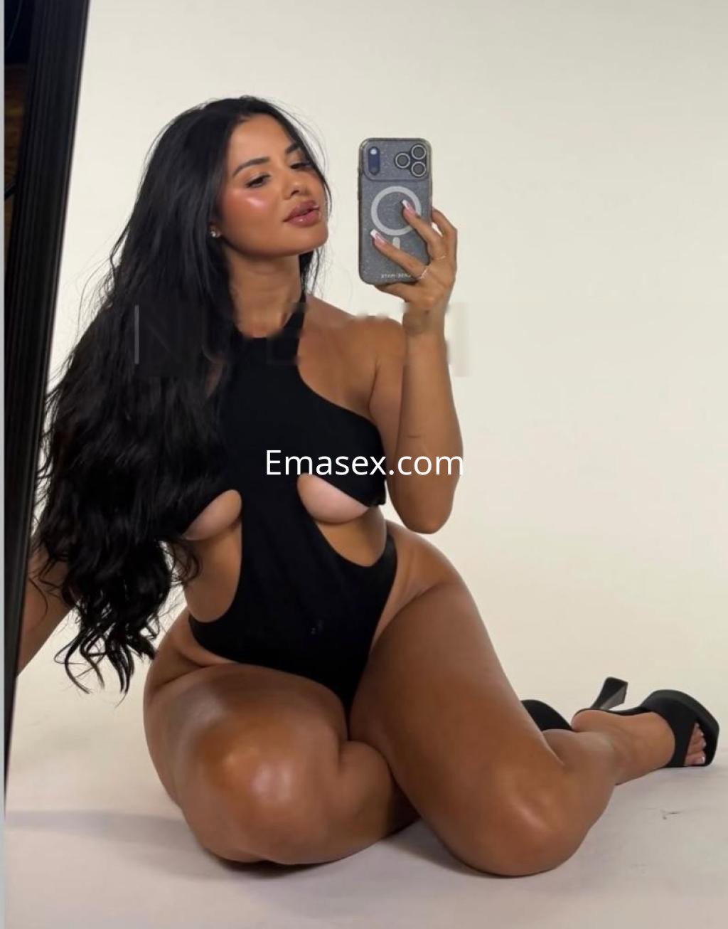 La escort de Barcelona posa en un body negro con cortes, sentada en el suelo y mirando hacia la cámara.