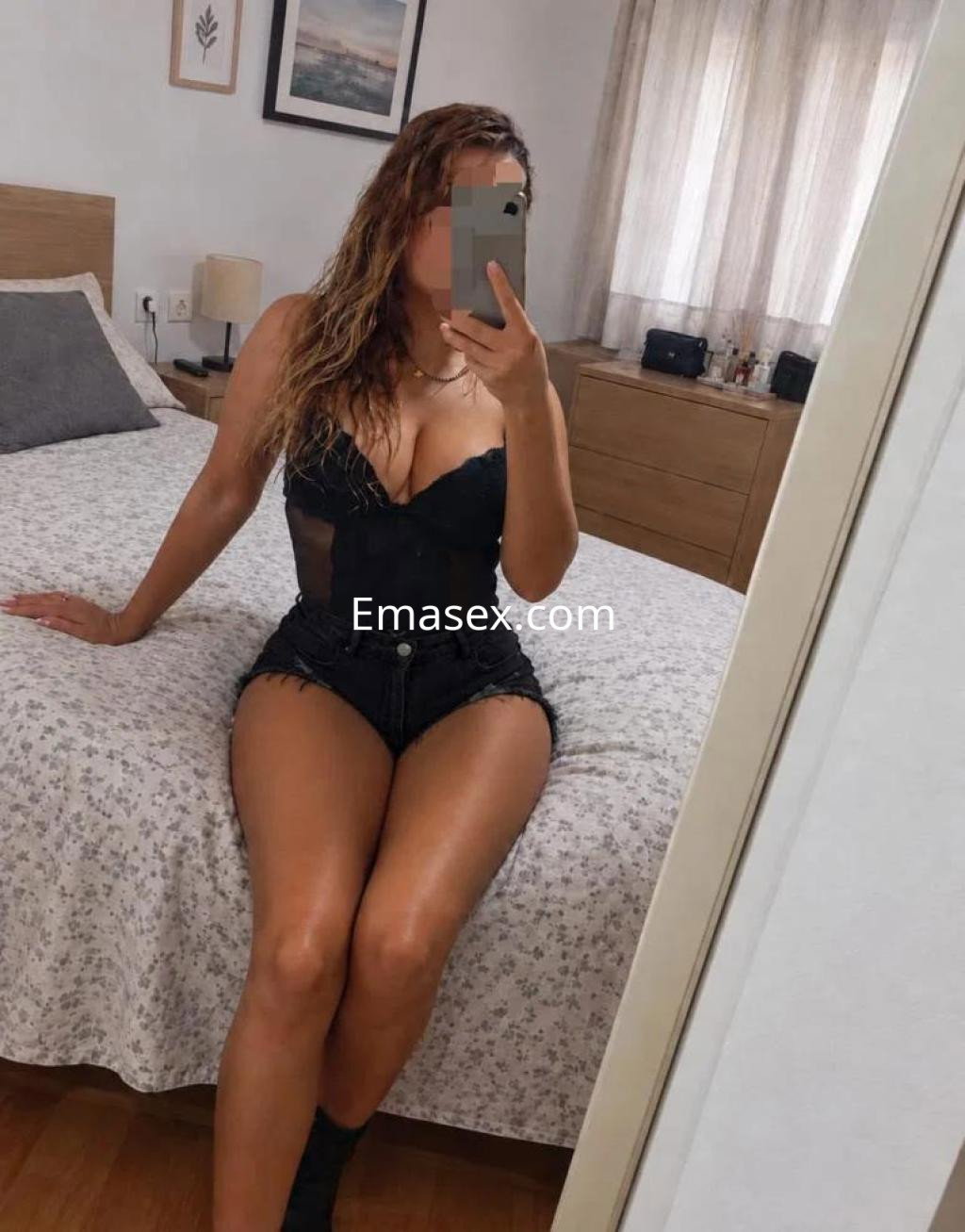 Escort en Alicante posando frente al espejo, con cabello ondulado y un atuendo negro que incluye un body y shorts.
