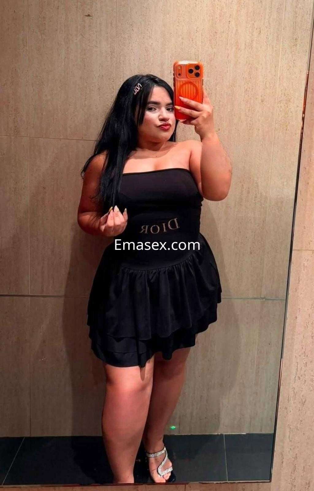 Escort en Murcia posando frente a un espejo con un vestido negro sin mangas y sandalias blancas, mostrando una sonrisa.