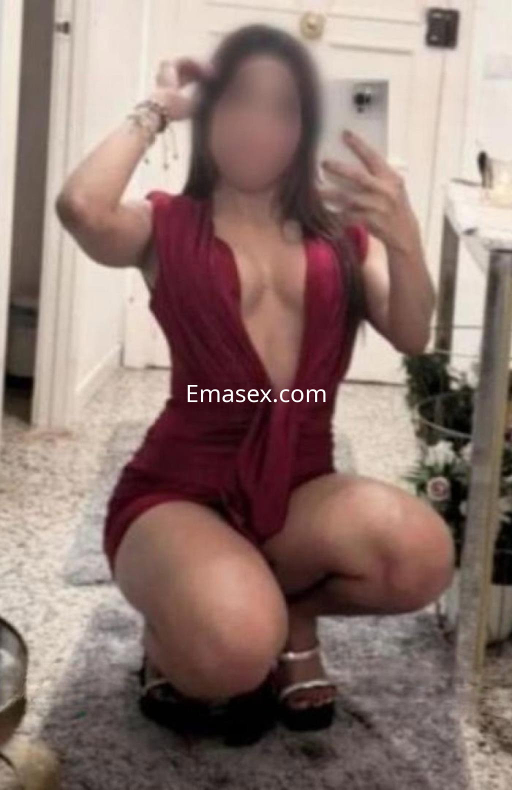 Escort en Zamora, agachada y posando frente al espejo, con un mono rojo de escote profundo y sandalias.