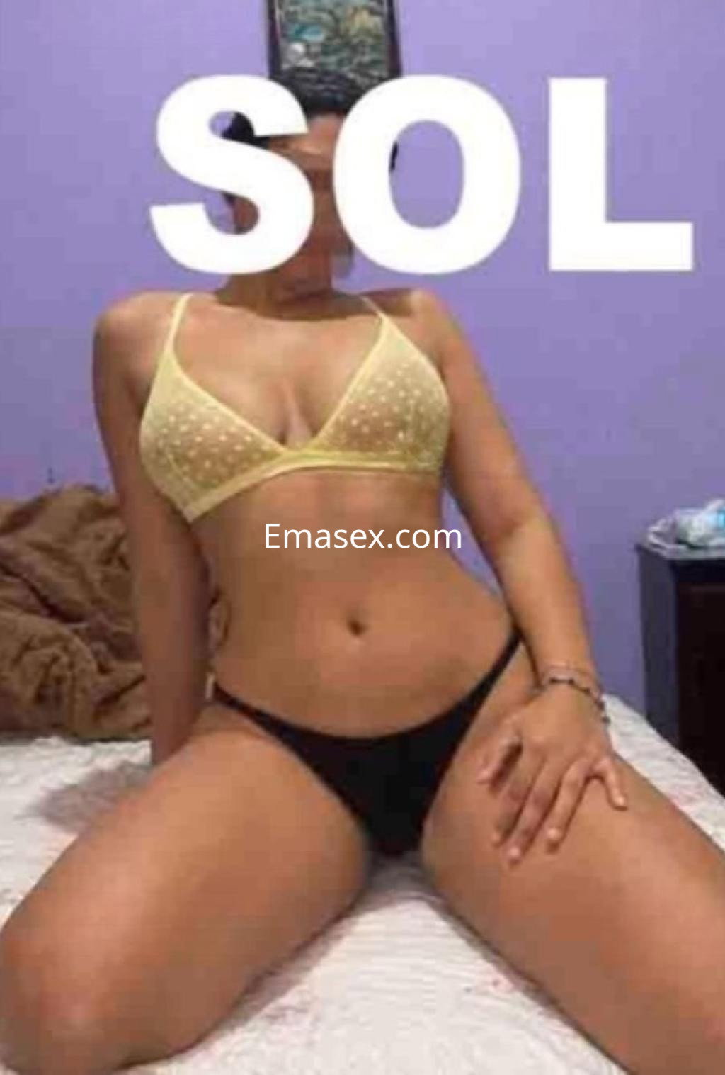 Puta en Valladolid se sienta en la cama, luciendo un sujetador amarillo y una tanga negra en un ambiente interior.
