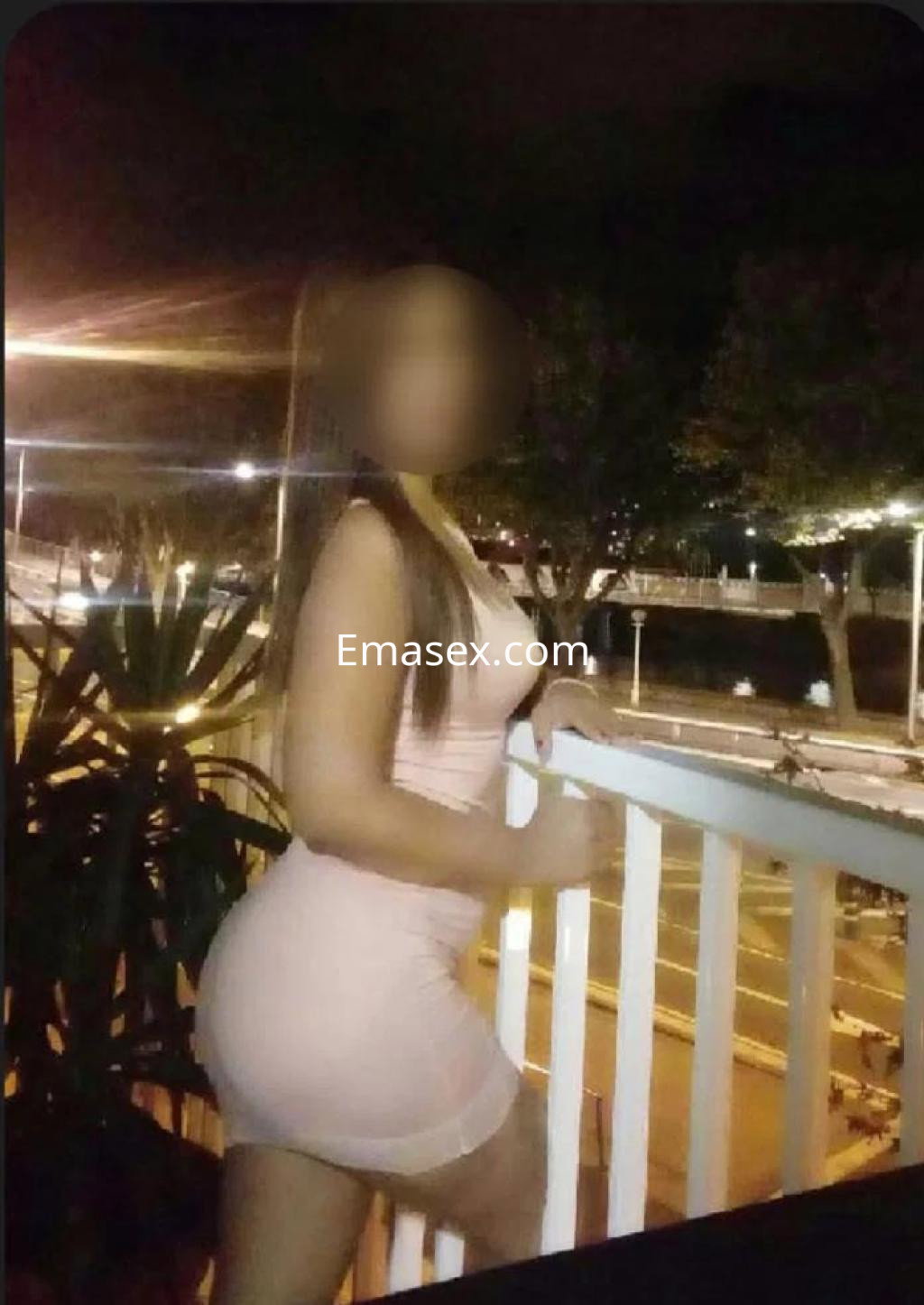 Escort en Huelva posando en un balcón, luciendo un vestido ajustado de color rosa en un ambiente nocturno.