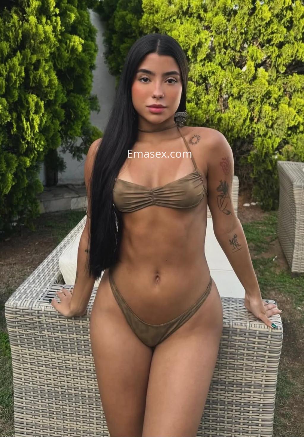 La escort luce un bikini marrón claro y posa de pie junto a un sofá en un entorno natural en Málaga.