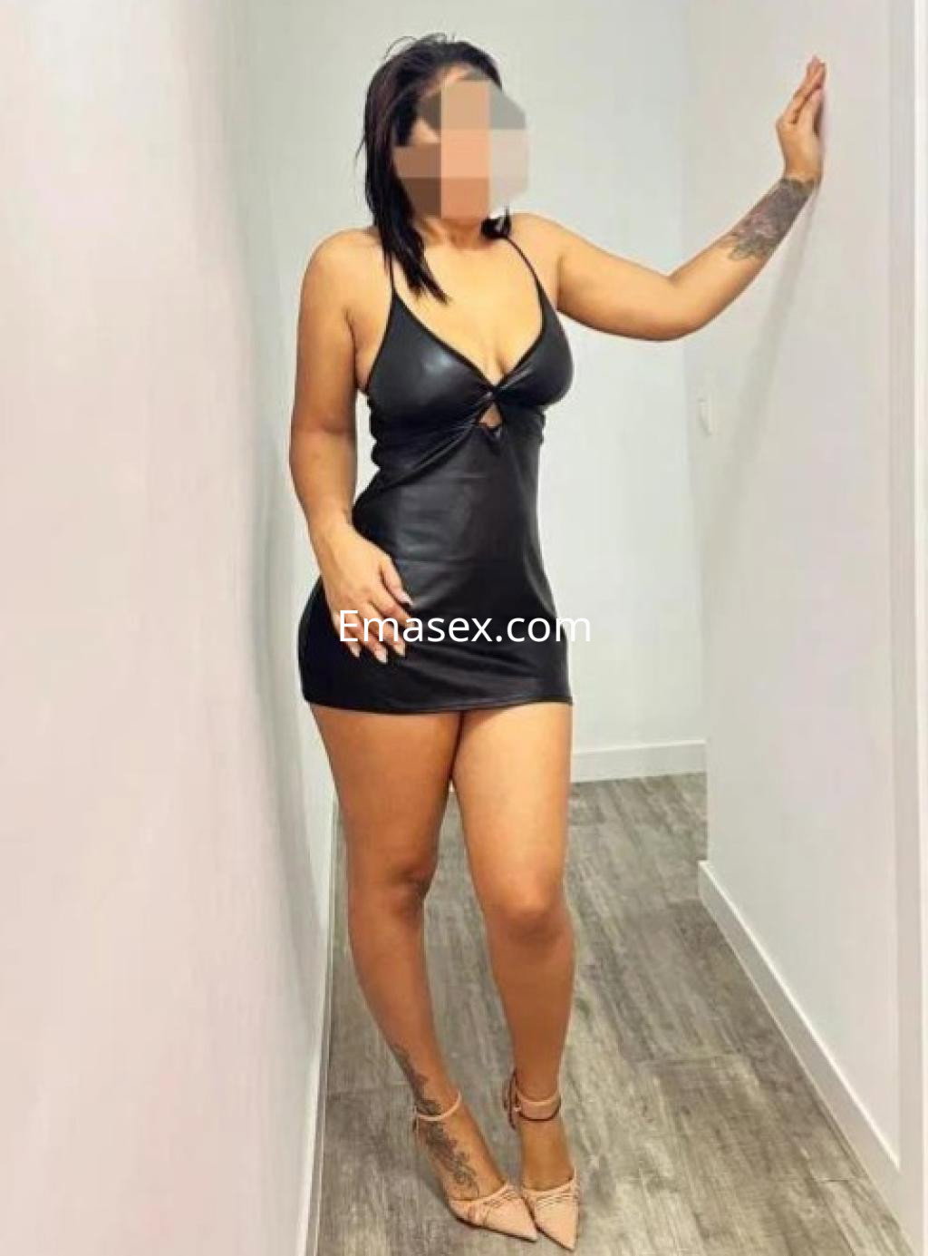 Puta en Burgos, luciendo un vestido negro ajustado y tacones, posando de pie junto a una pared blanca.