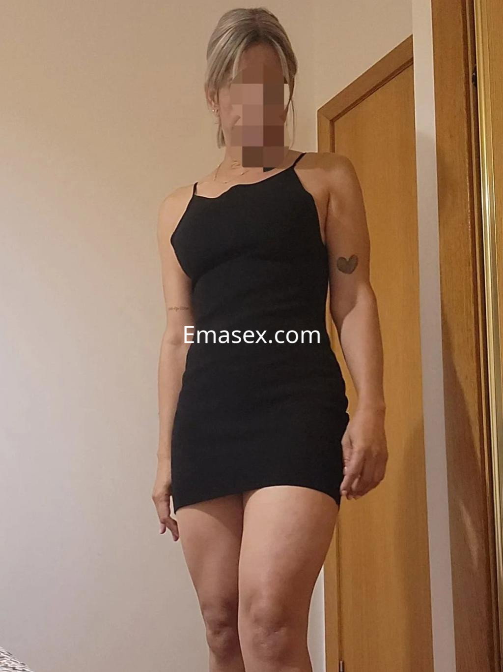 Escort en Córdoba viste un vestido negro ajustado, posando de pie en una habitación con paredes claras.