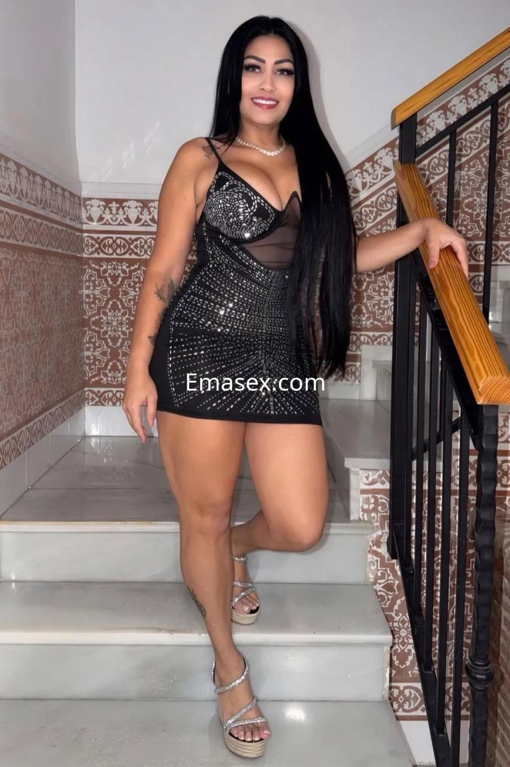 Puta en Córdoba con cabello largo y lacio, vistiendo un vestido corto negro con brillantes mientras posa en una escalera.