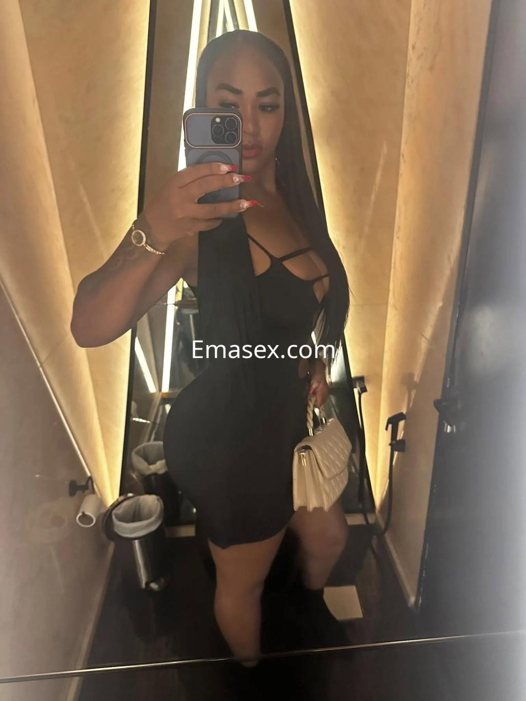 Escort en Zaragoza, posando frente al espejo con un vestido negro ajustado y bolso claro, mostrando una larga melena.