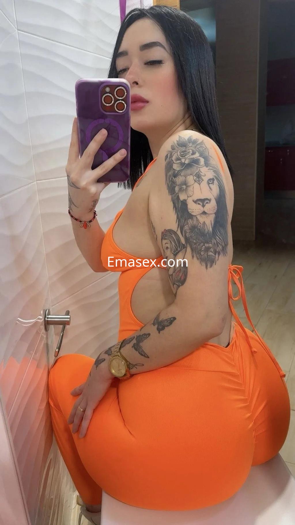 Puta en Santander posando en un traje naranja ajustado, mostrando tatuajes y un peinado lacio mientras se mira en el espejo.