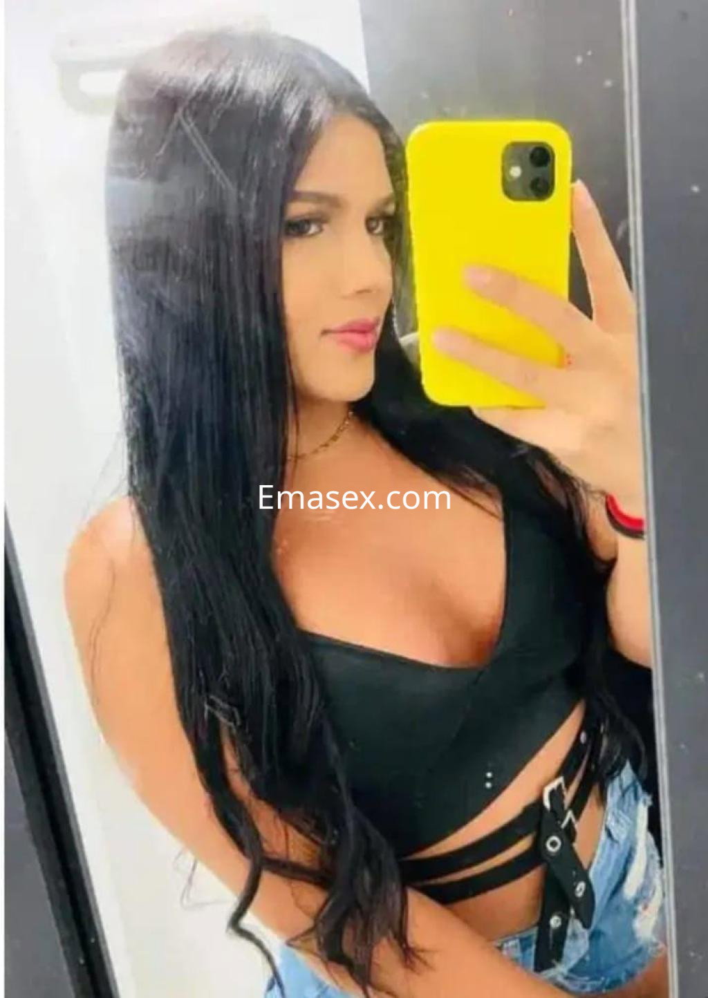 Escort trans en Pamplona con cabello largo y lacio, luciendo un top negro y jeans, posando frente al espejo.