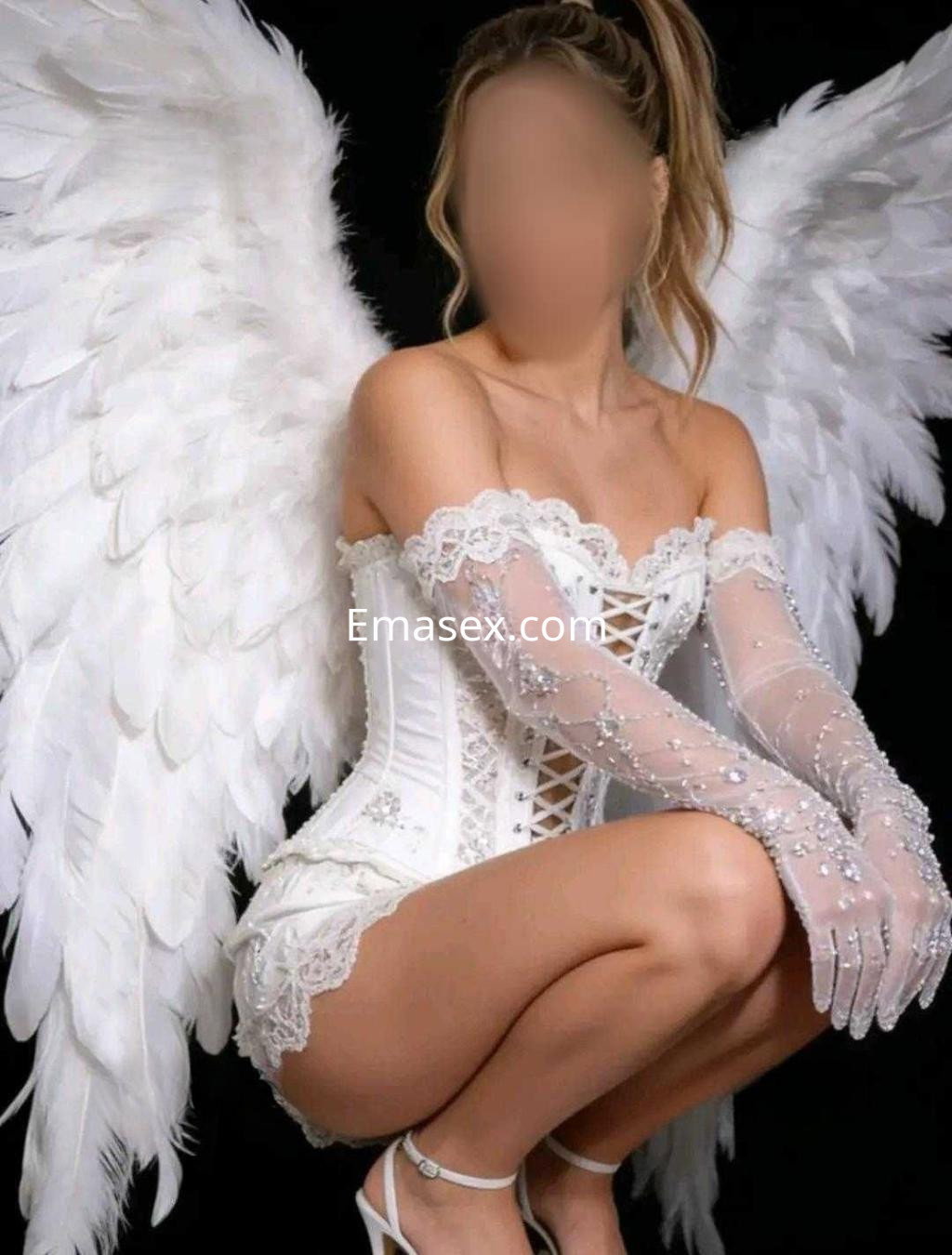 Escort en Alicante luciendo un corsé blanco con detalles brillantes y alas de plumas, en un fondo oscuro.