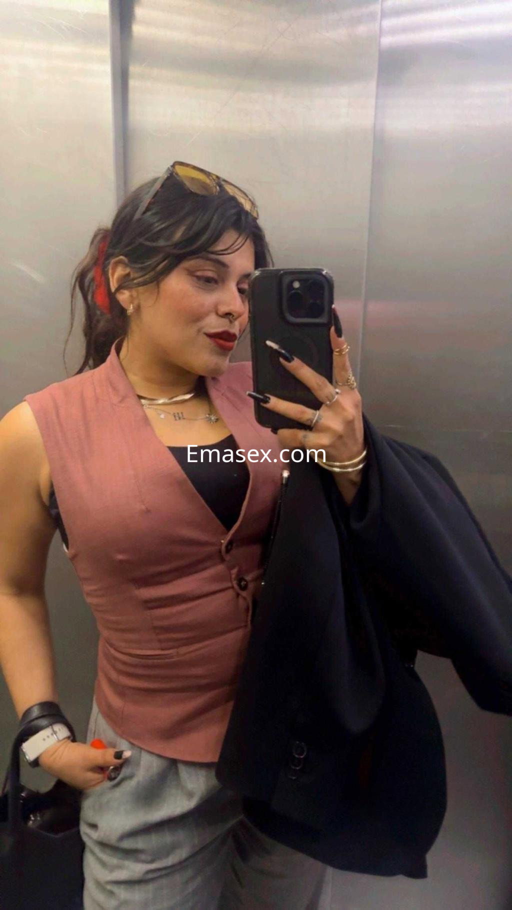 La puta posa en un ascensor con blusa rosa, pantalones claros y un abrigo negro, mostrando confianza.
