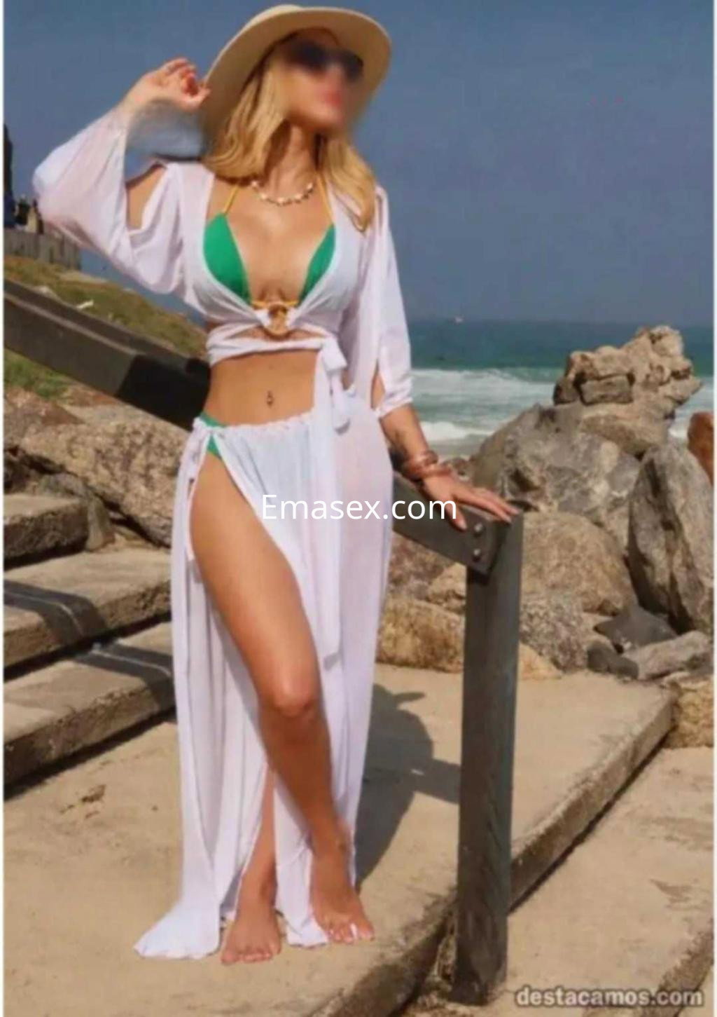 Escort en Badajoz con cabello rubio, usando un bikini verde y una túnica blanca en un entorno playero.