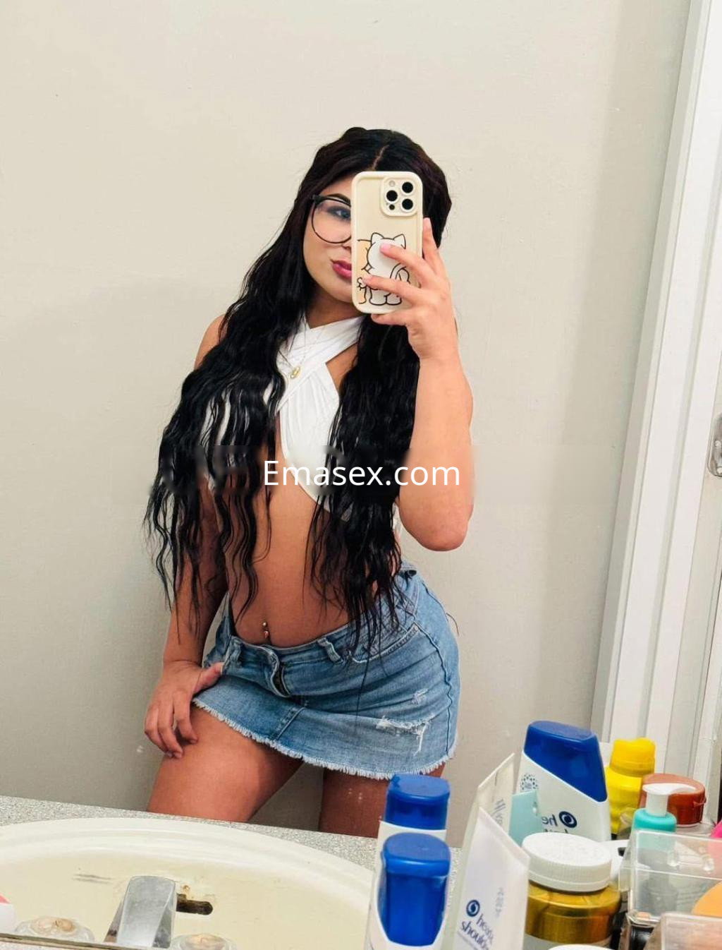 Escort en Alicante con cabello largo y oscuro, vistiendo un top blanco y una mini falda de mezclilla, posando frente al espejo.