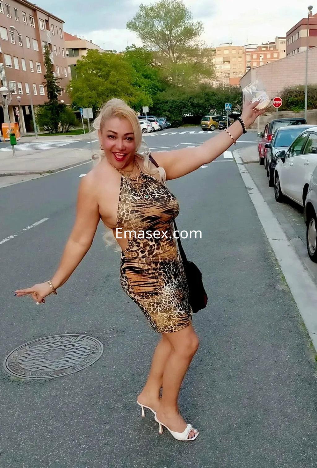 Escort en Zaragoza con vestido de estampado animal, sonriente y posando en una calle con coches aparcados.