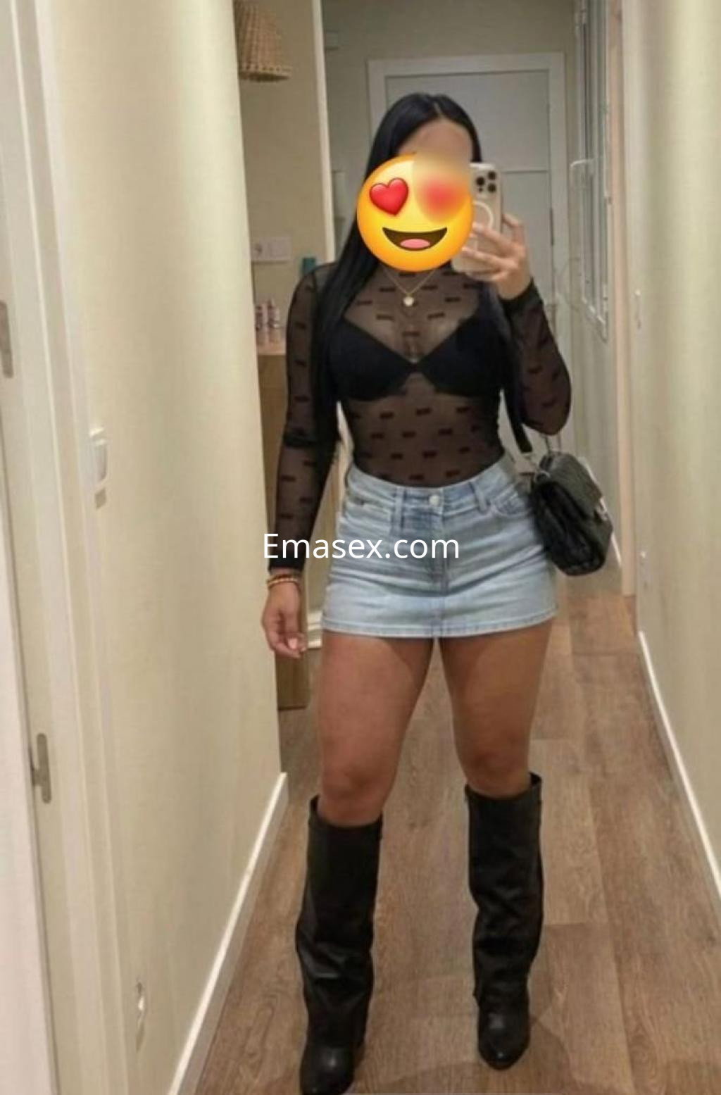 Puta en Granada luce un top negro transparente y una falda de mezclilla, posando en un pasillo moderno.