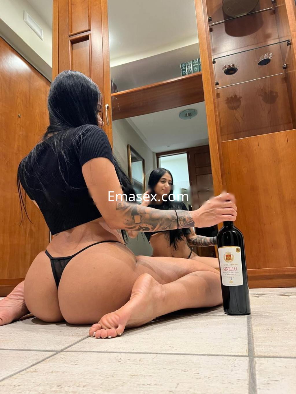 Escort en Girona sentada en el suelo, con un top negro y tanga, sosteniendo una botella de vino.
