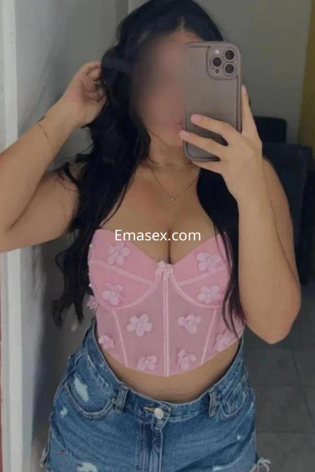 Escort de Córdoba con cabello largo y ondulado, vistiendo un corset rosa con flores y shorts de mezclilla, posando con un espejo.