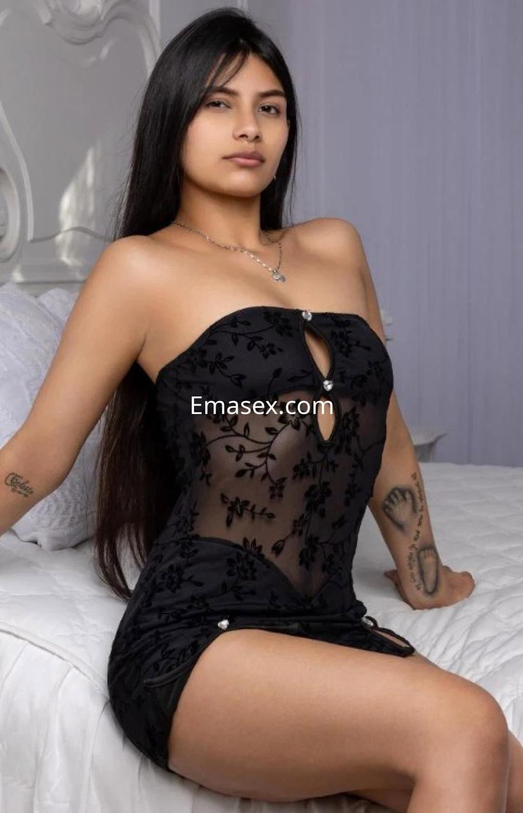 Escort en Castellón posando en una cama con un vestido negro ajustado y detalles florales, luciendo cabello largo.