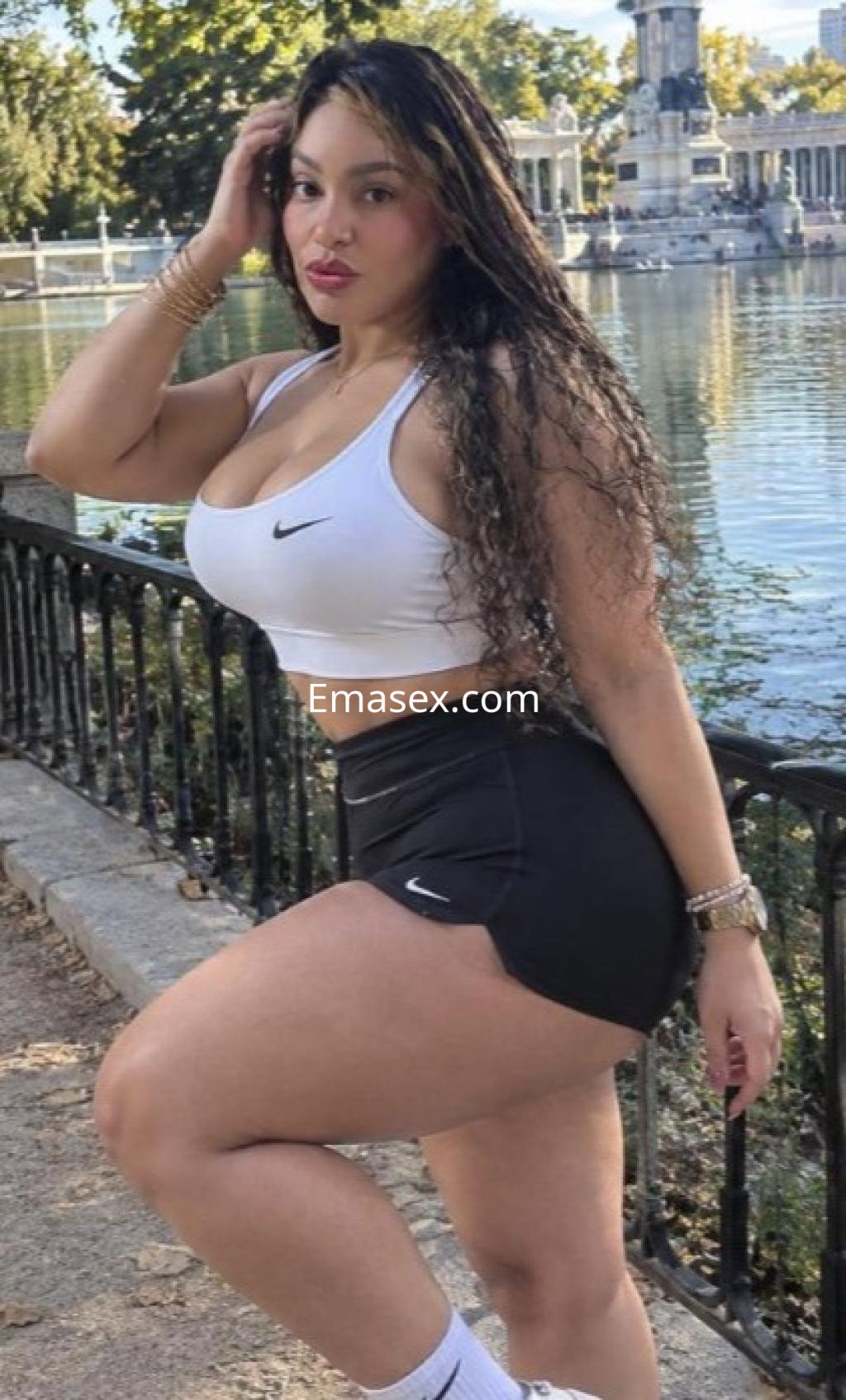 Puta posando junto a un lago en Castellón, viste un top blanco y una falda negra, con cabello largo y rizado.