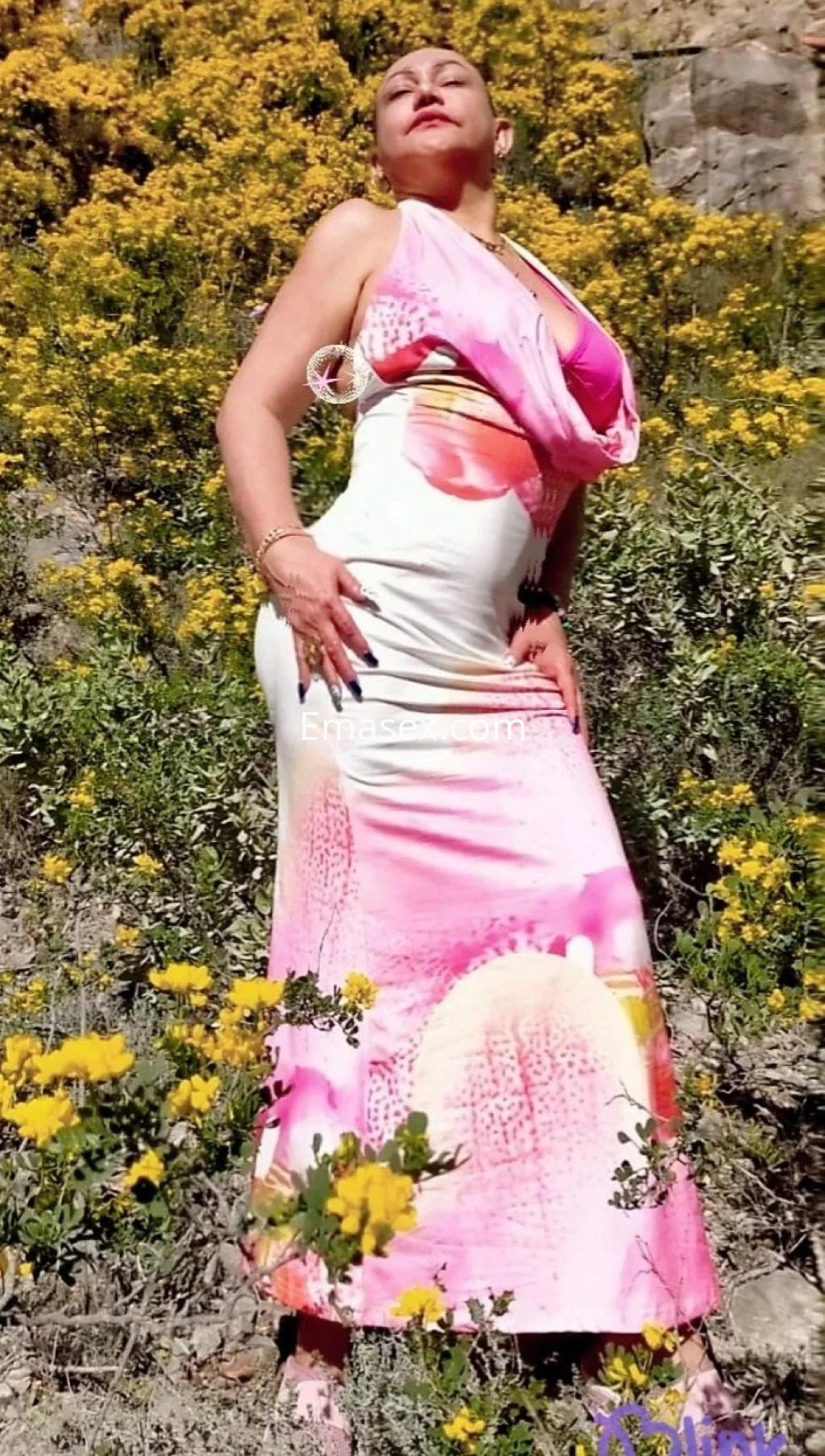 Escort en Zaragoza posando en un entorno natural, luciendo un vestido largo de colores suaves entre flores amarillas.