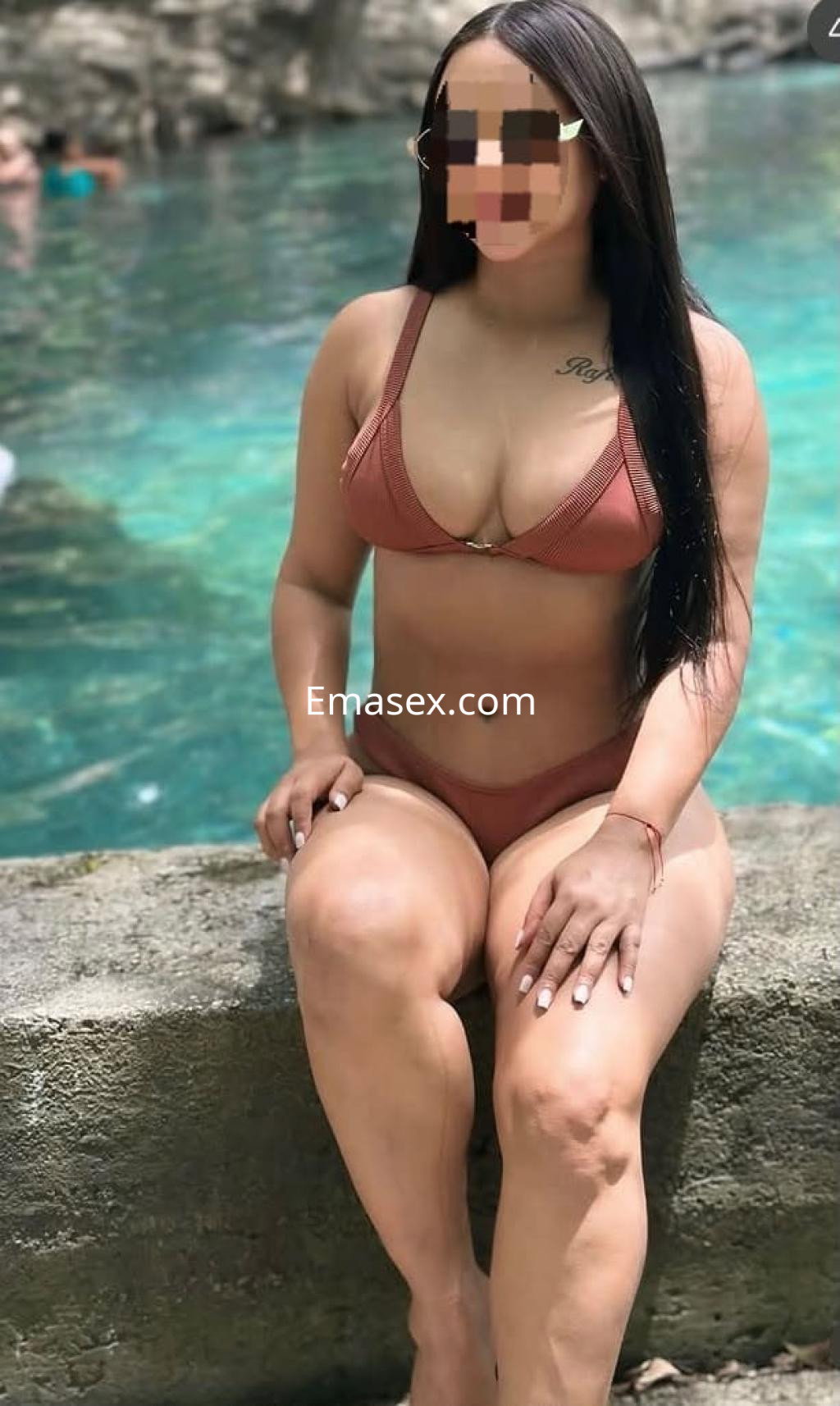 La puta en Huelva posa sentada junto a un agua cristalina, vistiendo un bikini marrón que destaca su figura.
