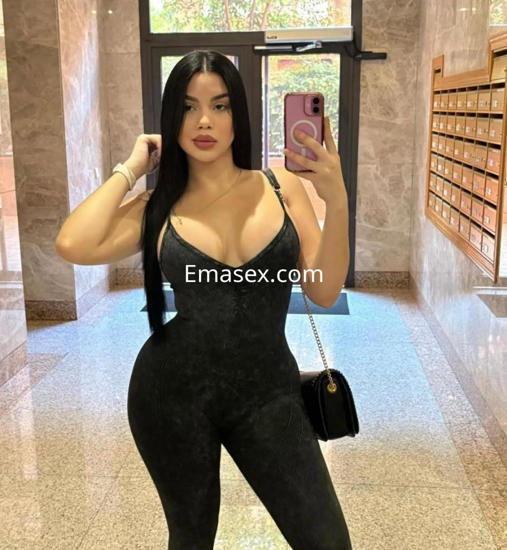Escort en Las Palmas con cabello lacio y oscuro, vistiendo un mono negro ajustado, posando en un vestíbulo.